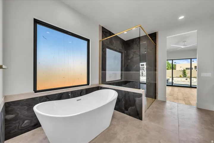 Freestanding soaking tub, large format tile flooring, black wall-mounted window, glass-enclosed shower with gold-finish trim, and dark-toned wall paneling