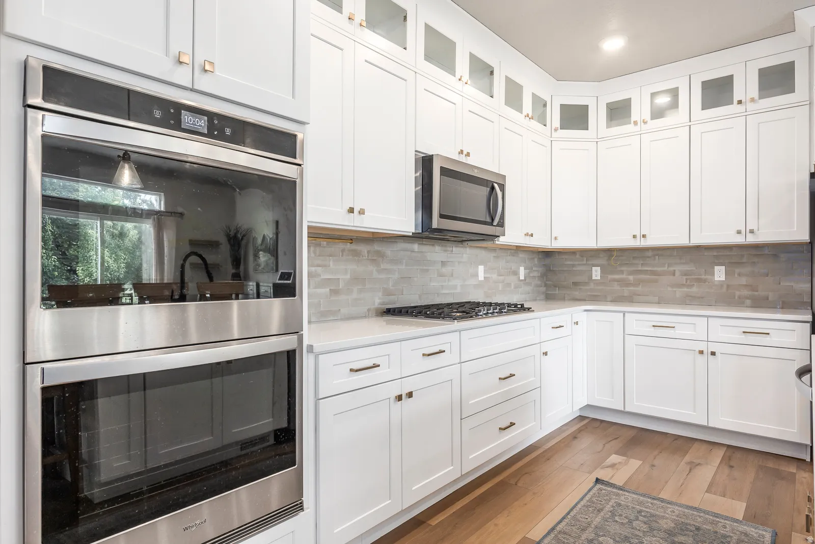 White shaker cabinetry, stone backsplash, built-in stainless steel double oven, stainless steel microwave, and wood-finish flooring