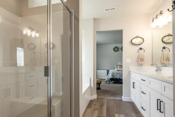 Walk-in shower with clear glass enclosure, dual vanity with white cabinetry, integrated sinks, chrome fixtures, and wood-finish flooring
