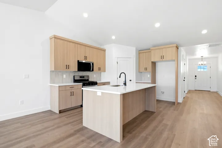 Kitchen featuring light wood-finish cabinetry, a central island with a sink and matte black faucet, stainless steel appliances, and wood-finish flooring