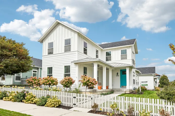 White exterior with board and batten and horizontal siding accents, dark roof, front porch with columns, a vibrant turquoise entry door, and a white picket fence