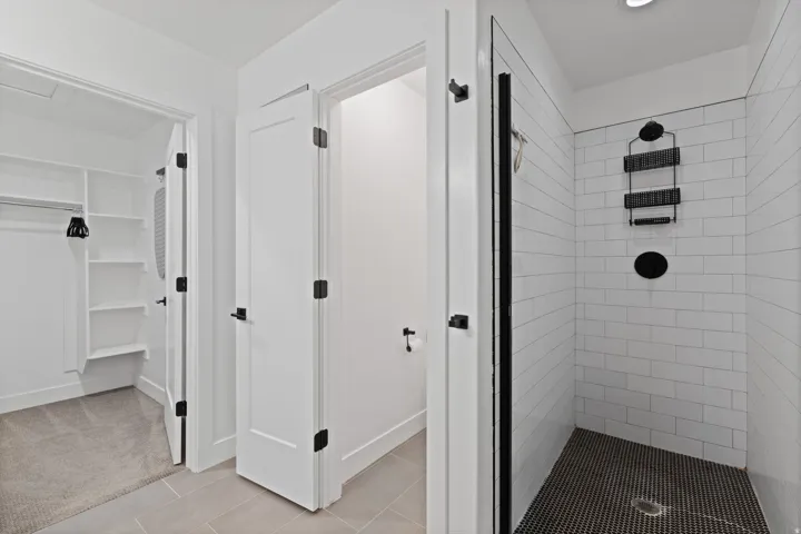Spacious walk-in closet with built-in shelving, white subway tile shower with dark grout, black penny tile shower floor, matte black shower fixtures, and light gray large format tile flooring