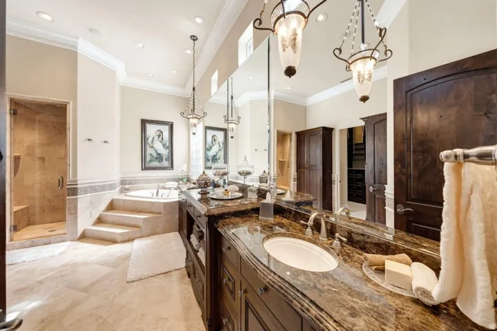 Dual vanity with stone countertop, built-in soaking tub with stone steps, glass-enclosed shower, dark wood cabinetry, and pendant light fixtures