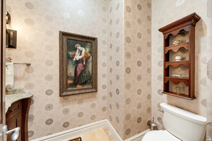 Decorative wall-mounted shelving unit, patterned wallpaper, wall sconce lighting, vanity with stone countertop, and wood-finish cabinetry