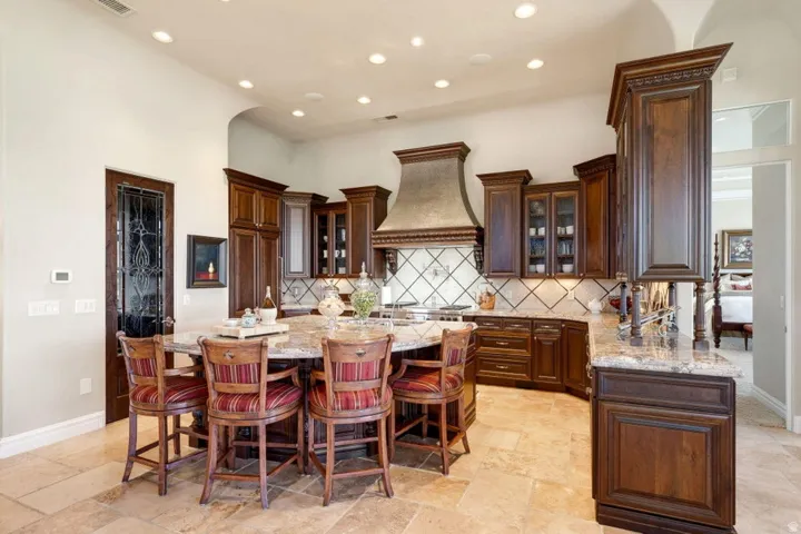 Expansive kitchen featuring a large central island with seating, dark wood cabinetry, light-toned tile flooring, and stone countertops