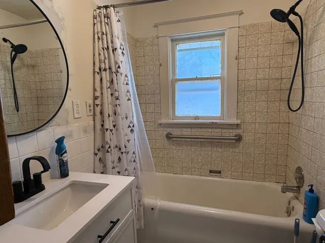 White vanity with a single basin sink and matte black faucet, full bathtub with a shower fixture, and a wall-mounted shower head
