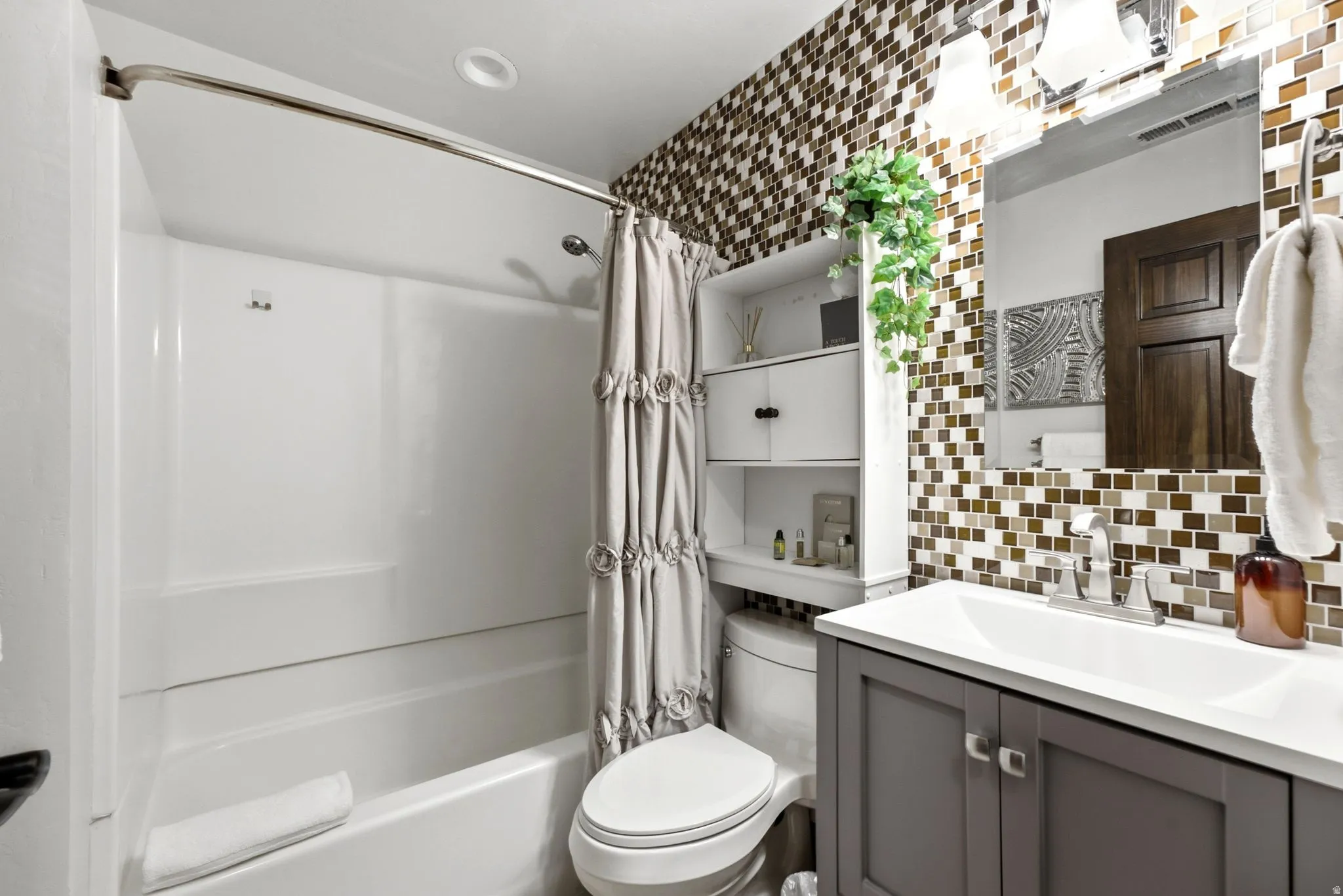 Full-height mosaic tile accent wall, white vanity with integrated sink, contemporary vanity light fixture, built-in shelving, and tub-shower combination