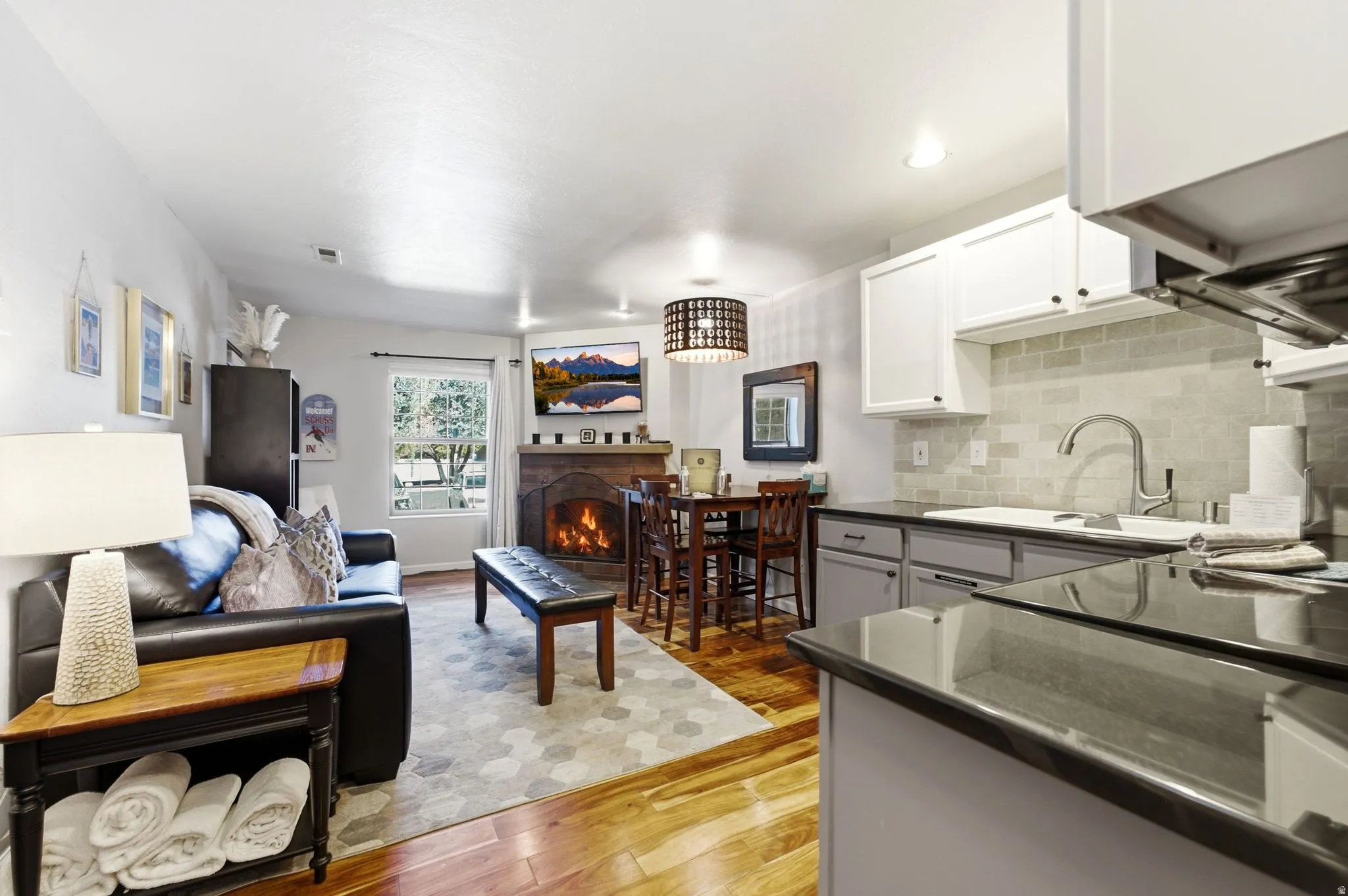 Wood-finish flooring, built-in fireplace, kitchen with white upper cabinetry and gray lower cabinetry, gray tile backsplash, and dark countertops