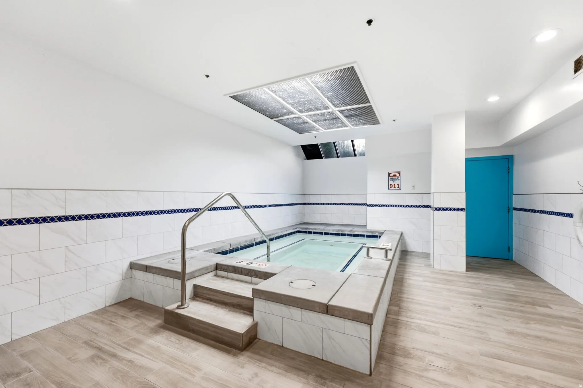 Indoor spa tub featuring a stainless steel handrail, light wood-finish flooring, and white tiled walls with a blue decorative accent