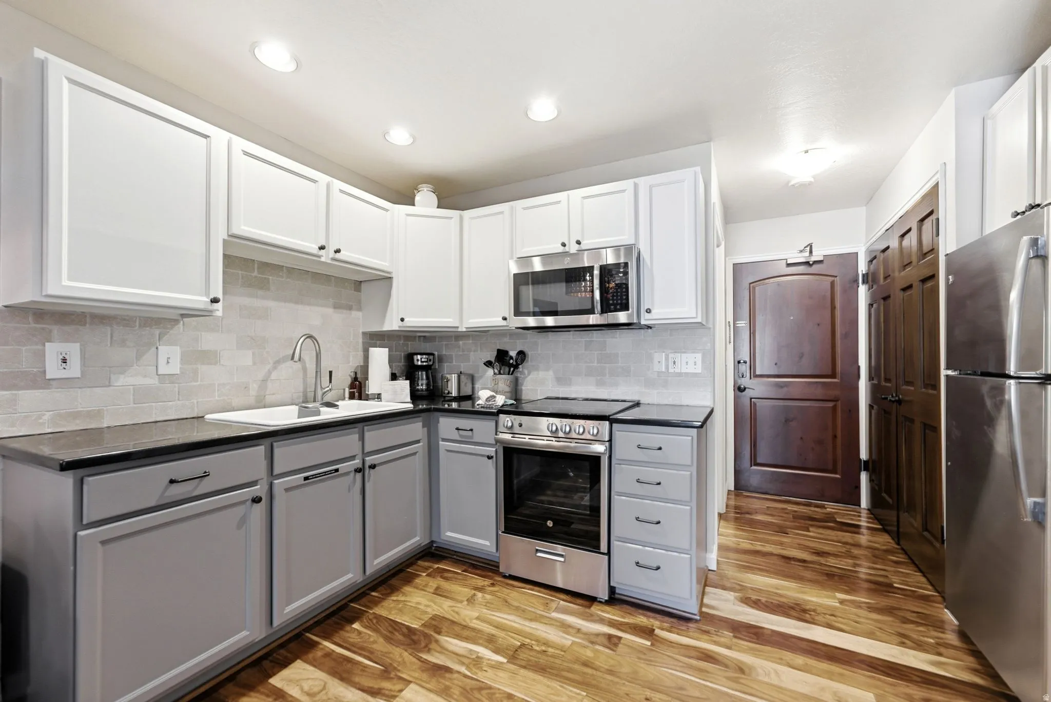 Contemporary kitchen featuring wood-finish flooring, two-tone cabinetry with black hardware, stainless steel appliances, dark countertops, and a tiled backsplash