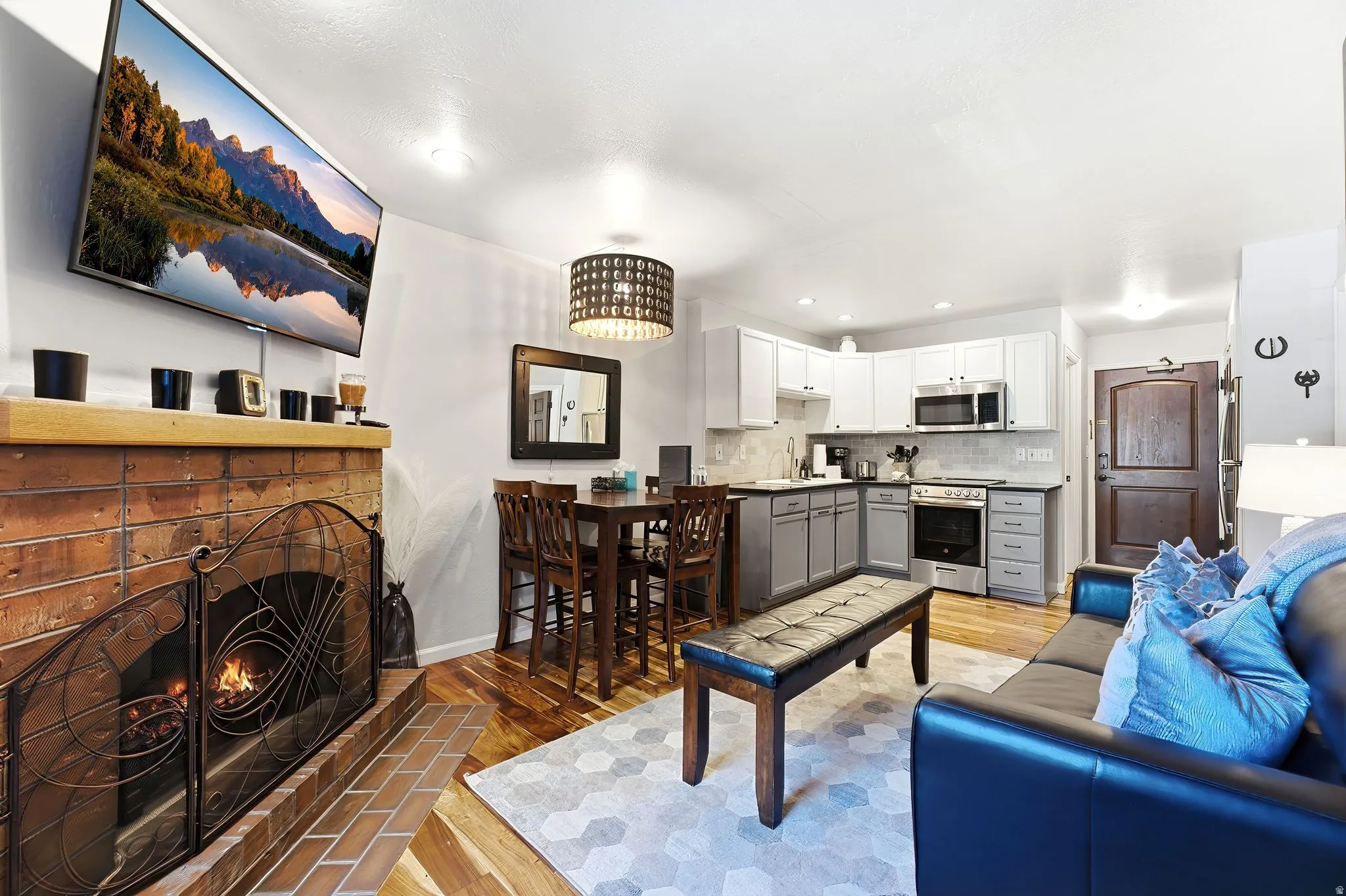 Brick fireplace with a metal screen, wood-finish flooring, kitchen with stainless steel appliances, two-tone cabinetry, and a subway tile backsplash