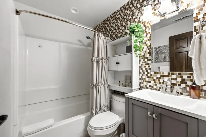 Full-height mosaic tile accent wall, white vanity with integrated sink, contemporary vanity light fixture, built-in shelving, and tub-shower combination