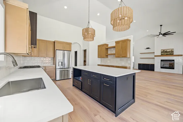 Vaulted ceilings with decorative light fixtures, kitchen island with dark cabinetry and light countertops, wood-finish flooring, stainless steel refrigerator, and a fireplace with a white tile surround