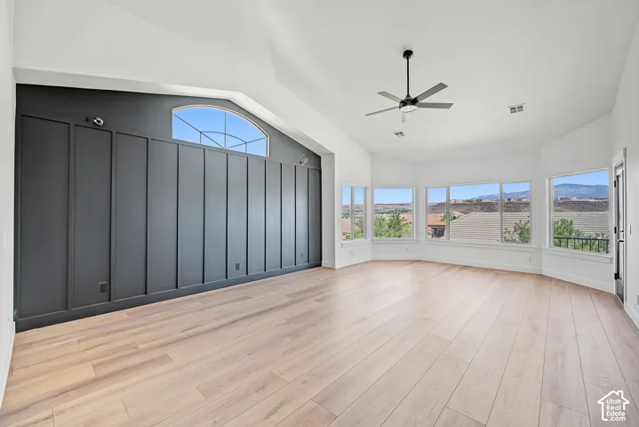 Vaulted ceiling featuring a contemporary ceiling fan, wood-finish flooring, accent wall with panel detailing, arched transom window, and a series of large windows
