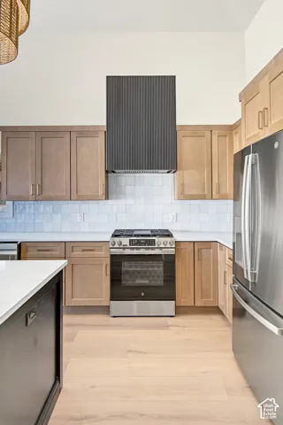 Vaulted ceiling kitchen featuring light wood-finish cabinetry, stainless steel appliances, a range with a dark textured hood, and light wood-finish flooring