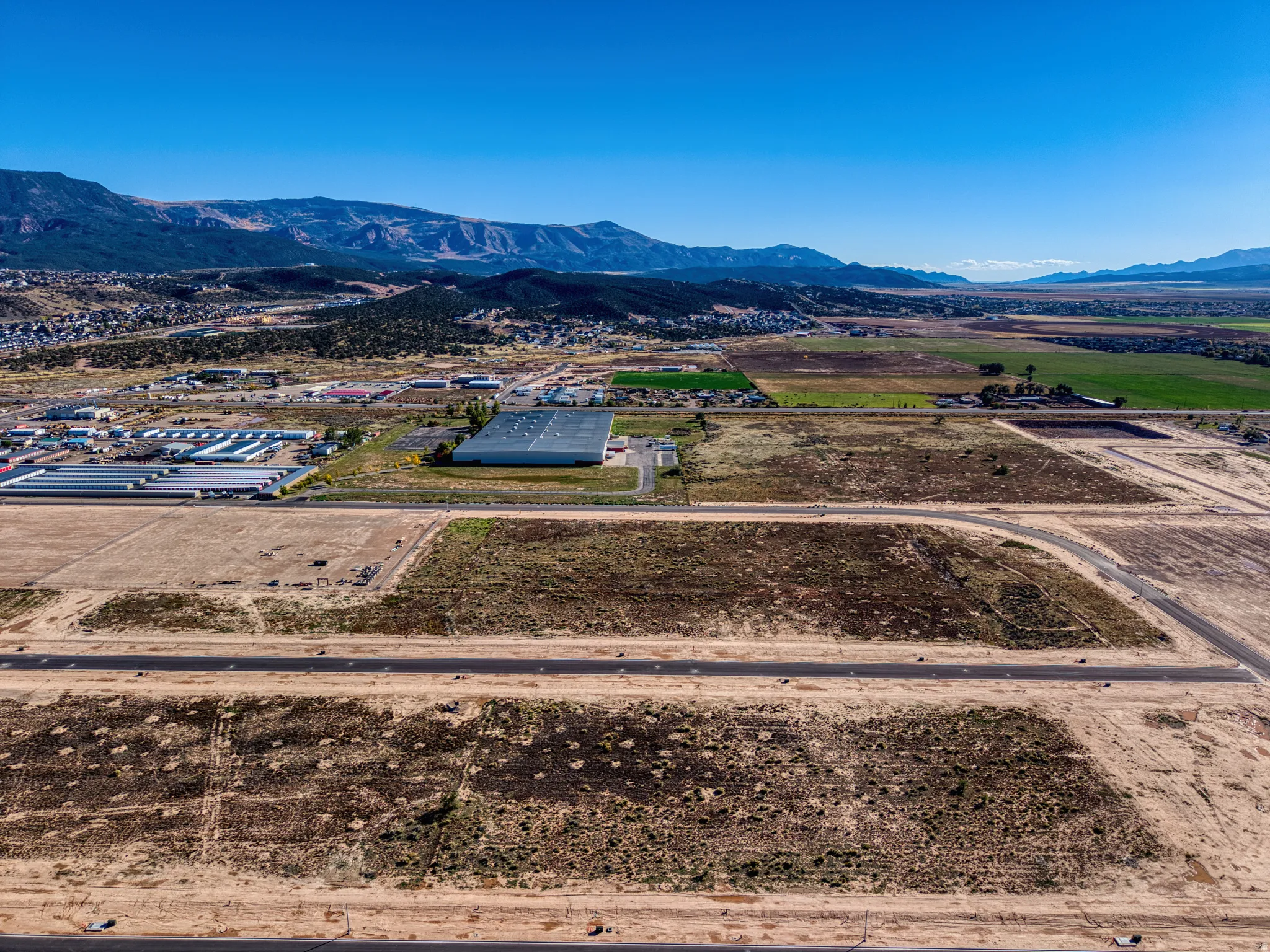 Expansive undeveloped land with visible graded areas, surrounding commercial structures, established roads, and distant mountainous terrain