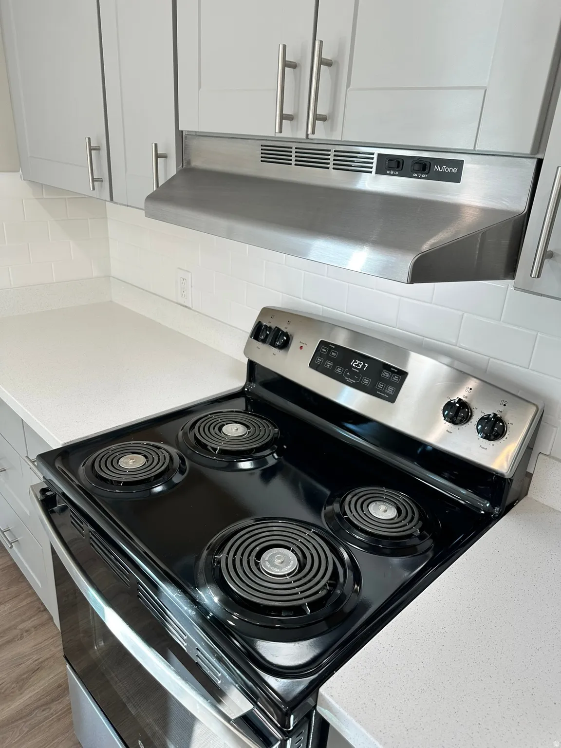 Stainless steel range hood, white subway tile backsplash, light-toned countertops, and wood-finish flooring