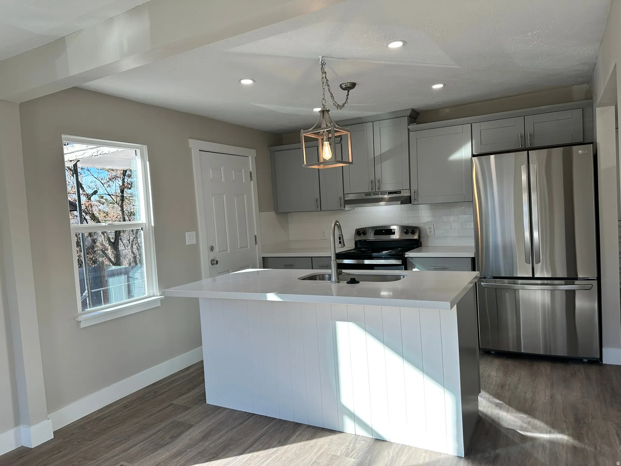 Kitchen featuring a central island with white paneling and a light-toned countertop, stainless steel appliances, grey cabinetry, white subway tile backsplash, and wood-finish flooring