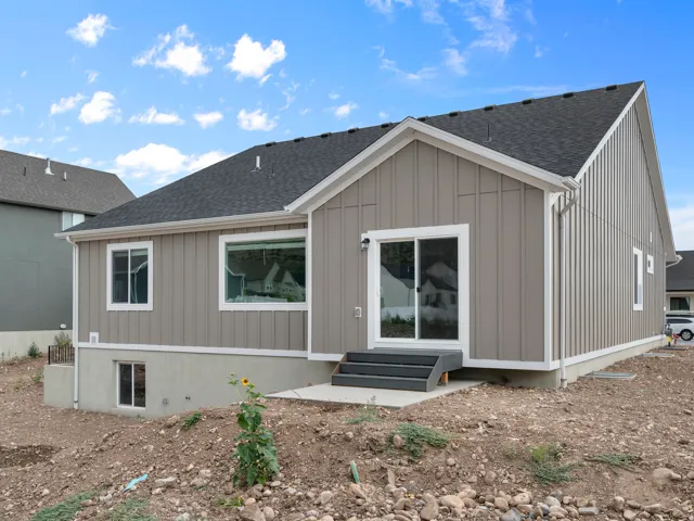 Board and batten siding, dark shingle roof, white trim accents, sliding glass door, and a concrete patio