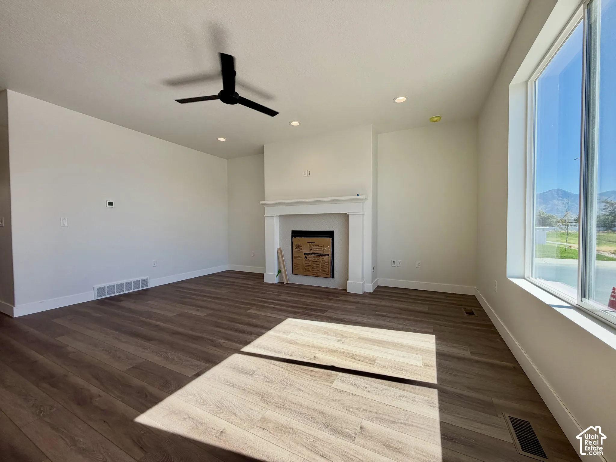Spacious room featuring wood-finish flooring, a contemporary ceiling fan, recessed lighting, a fireplace with a white mantel, and large windows