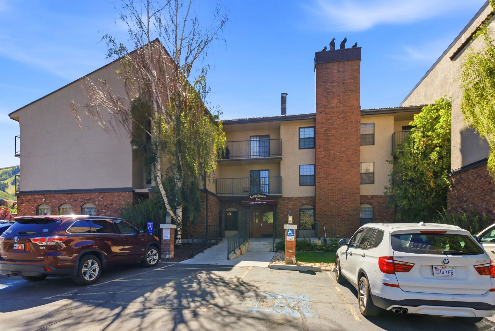 Multi-story building with brick veneer and stucco exterior, featuring multiple balconies with metal railings, a prominent brick chimney, and mature trees