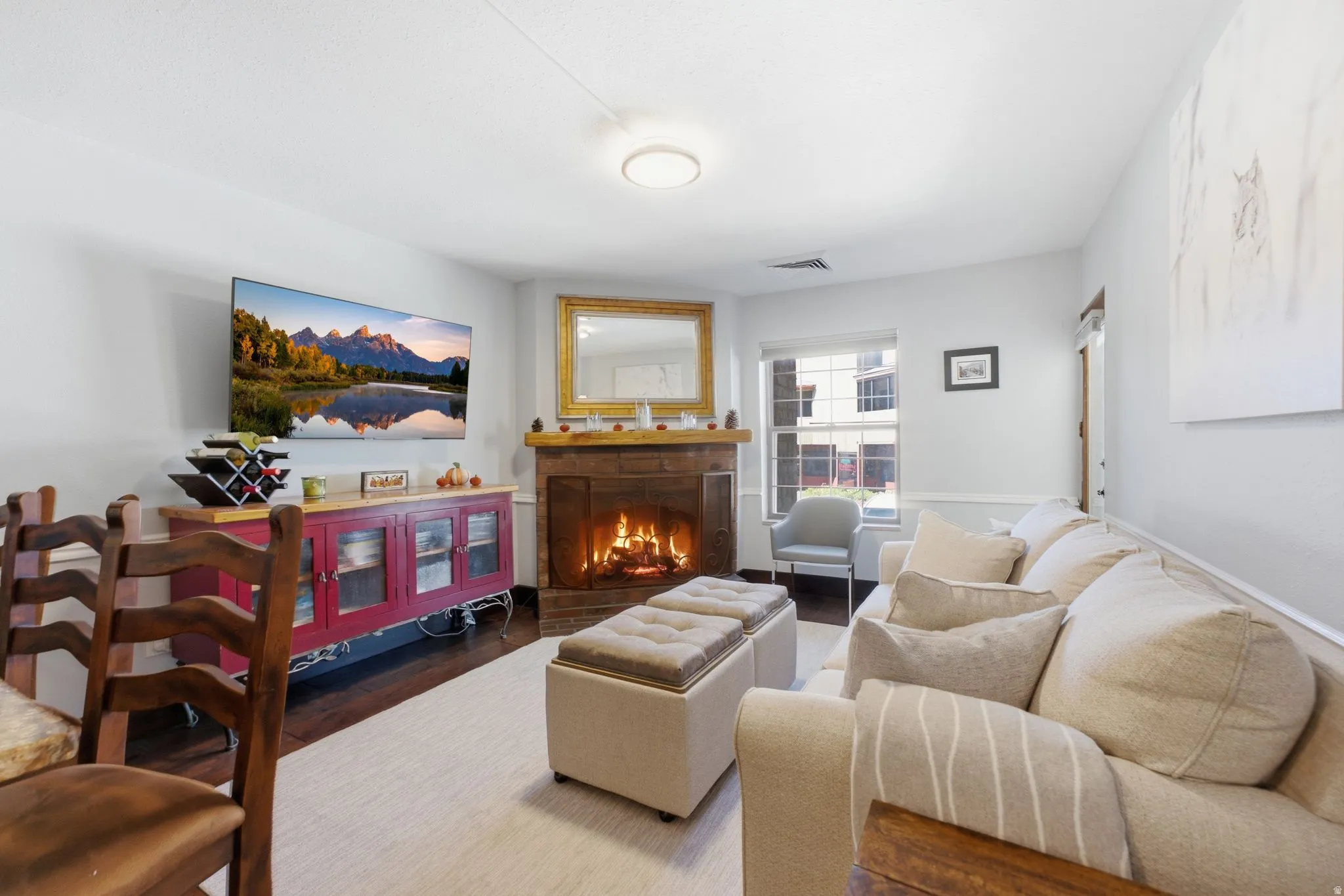 Wood-finish flooring with a light-toned area rug, a brick fireplace with wood mantel, and large windows with blinds