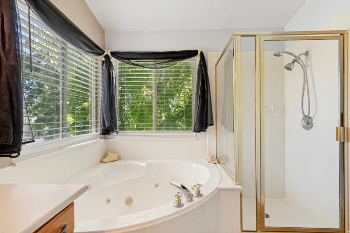 Corner soaking tub with integrated jets, multiple windows with blinds, a separate enclosed shower with a dual showerhead, and a vanity with a wood-finish cabinet