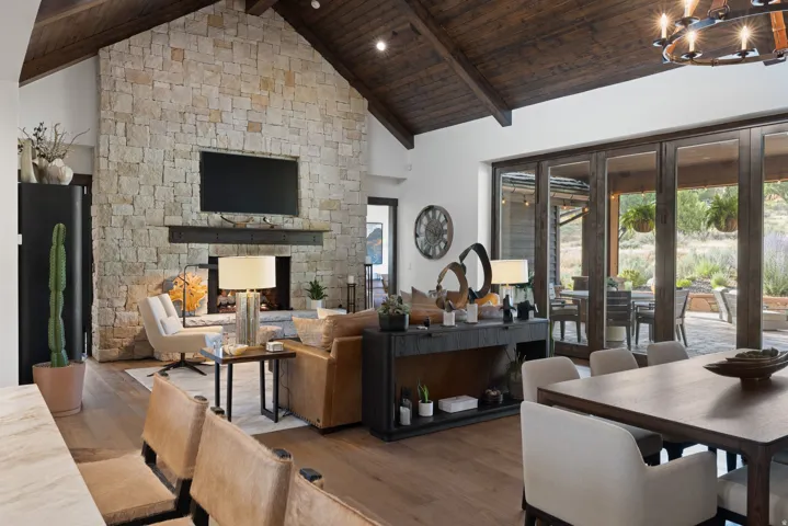 Vaulted wood-beamed ceiling, stone-clad fireplace with wood mantle, wide plank wood-finish flooring, and folding glass doors