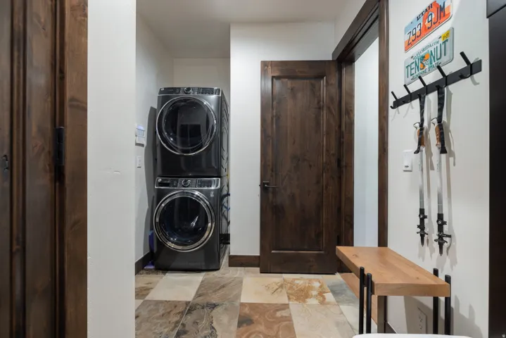 Stacked front-load washer and dryer, wood-finish interior door, multi-toned tile flooring, and a built-in wooden bench with a metal frame