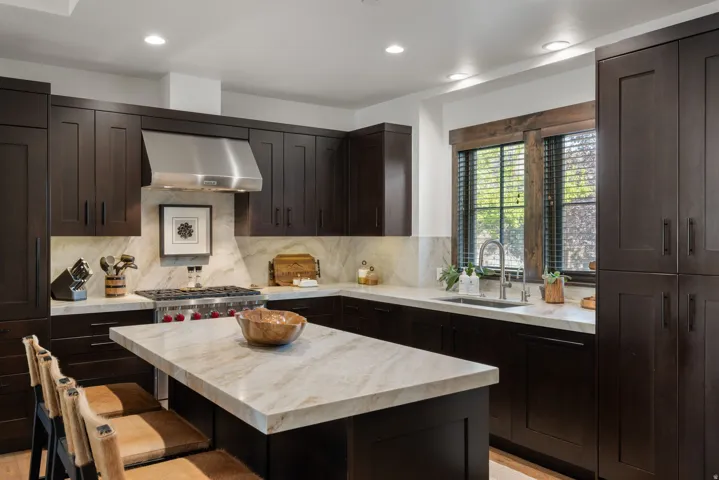 Dark wood-finish cabinetry with recessed panel doors, light-toned stone countertops, integrated stainless steel appliances, stainless steel range hood, and a built-in kitchen island