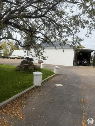 Exterior of an outbuilding with white metal siding, a dark metal roof, and an attached carport, featuring paved driveway access and masonry gate posts