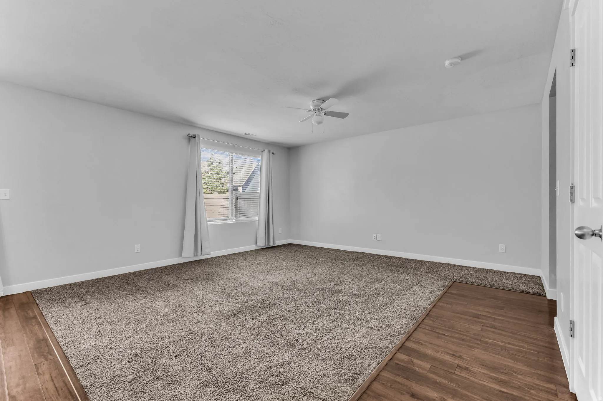 Spacious room featuring neutral carpeting, wood-finish flooring, a window with blinds, and a ceiling fan