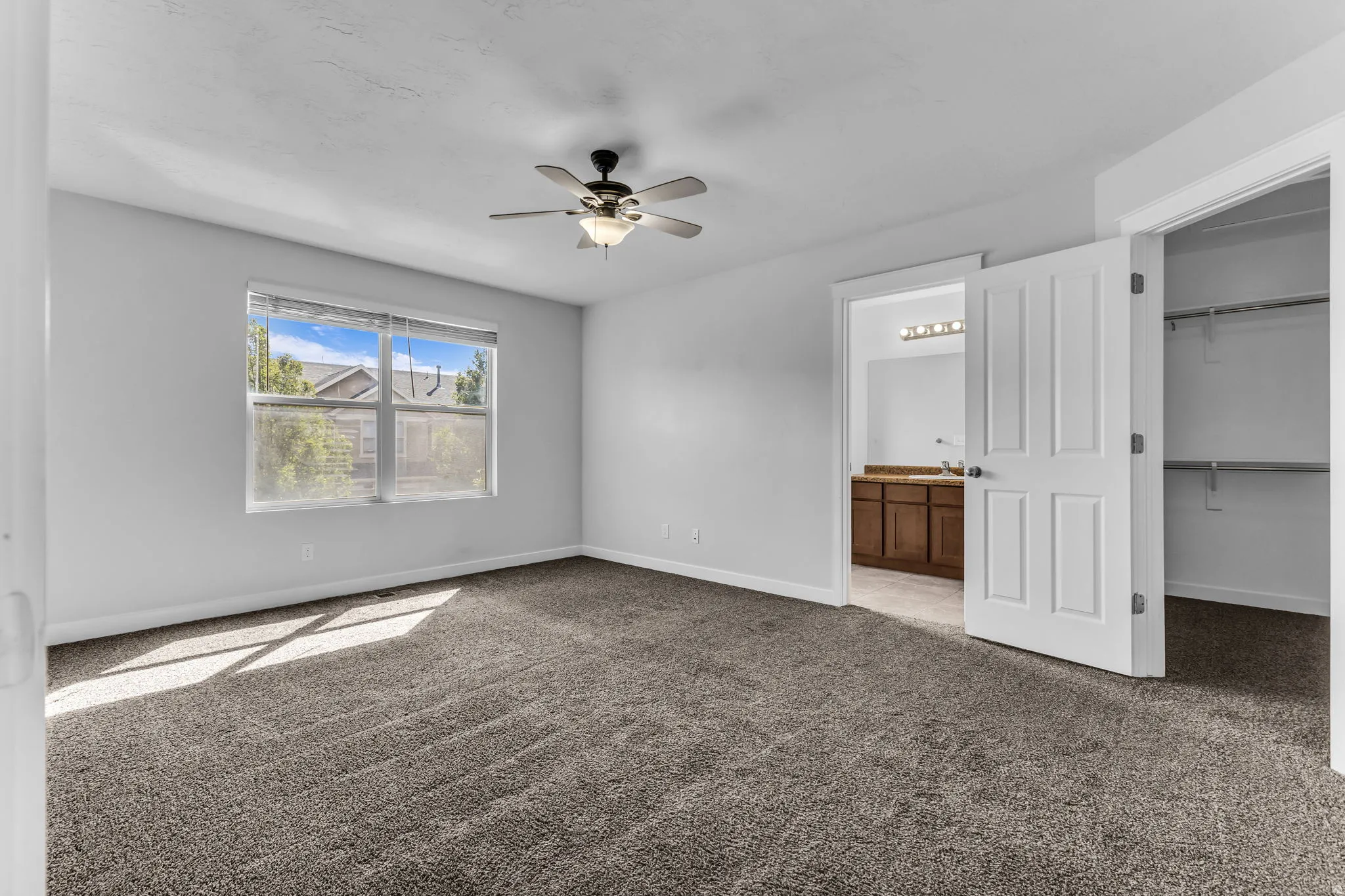 Neutral-toned carpet flooring, window with blinds, ceiling fan with integrated lighting, open doorway to a bathroom with a wood-finish vanity, and an open closet with shelving and hanging rods