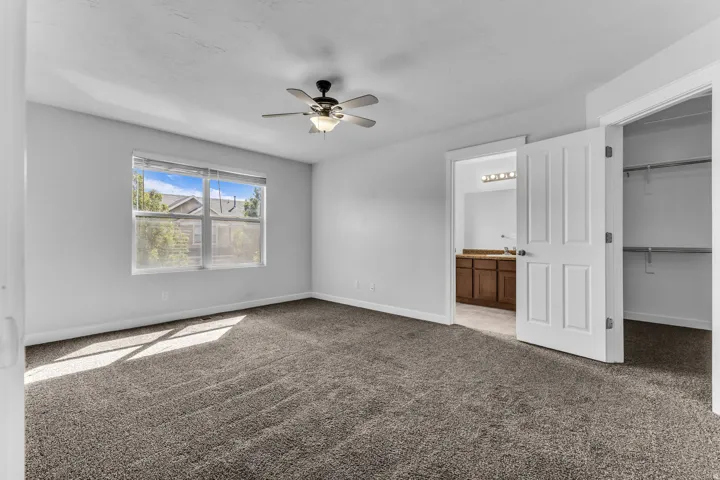 Neutral-toned carpet flooring, window with blinds, ceiling fan with integrated lighting, open doorway to a bathroom with a wood-finish vanity, and an open closet with shelving and hanging rods