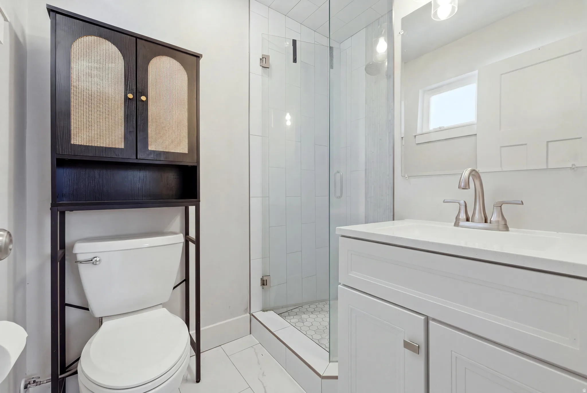 Walk-in shower with vertical tile surround and hexagonal floor, single vanity with white cabinetry and integrated sink, brushed nickel faucet, and dark wood-finish over-the-toilet shelving unit