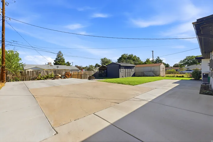 Expansive concrete driveway and patio area, featuring a manicured lawn, wood fencing, and two detached storage sheds