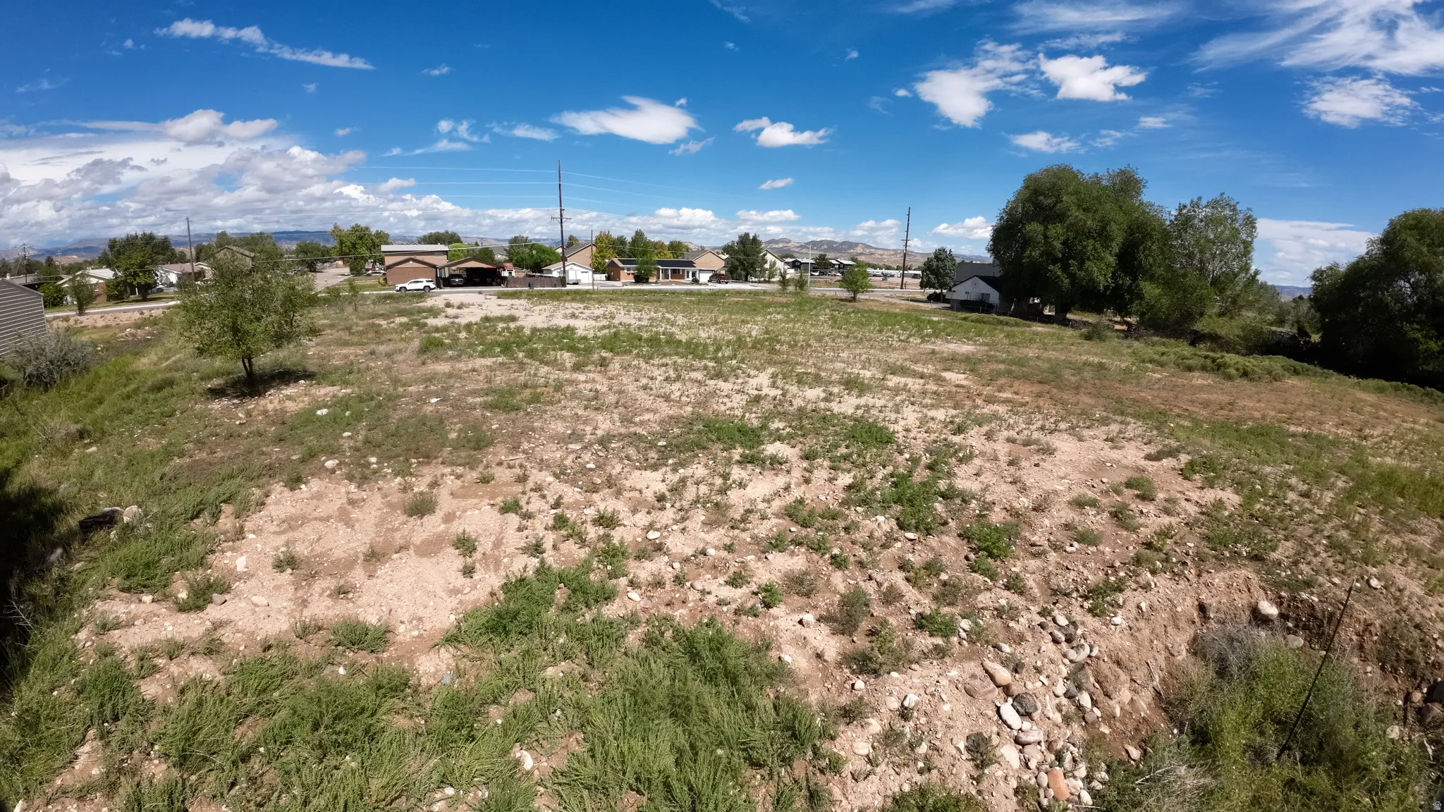 Expansive undeveloped lot featuring natural vegetation, scattered rocks, and distant residential structures
