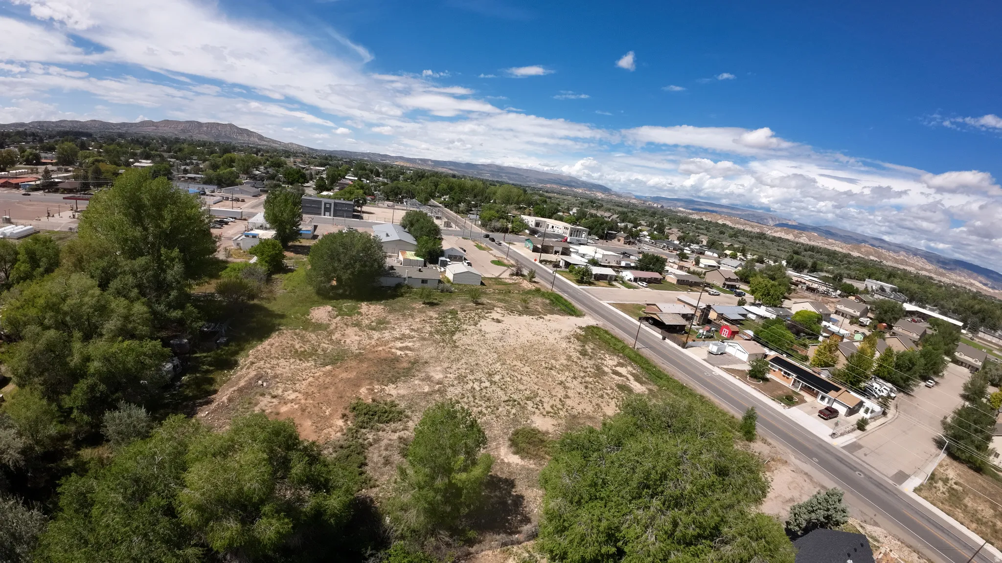 Expansive land parcel with surrounding residential and commercial structures, paved road frontage, and distant mountain backdrops