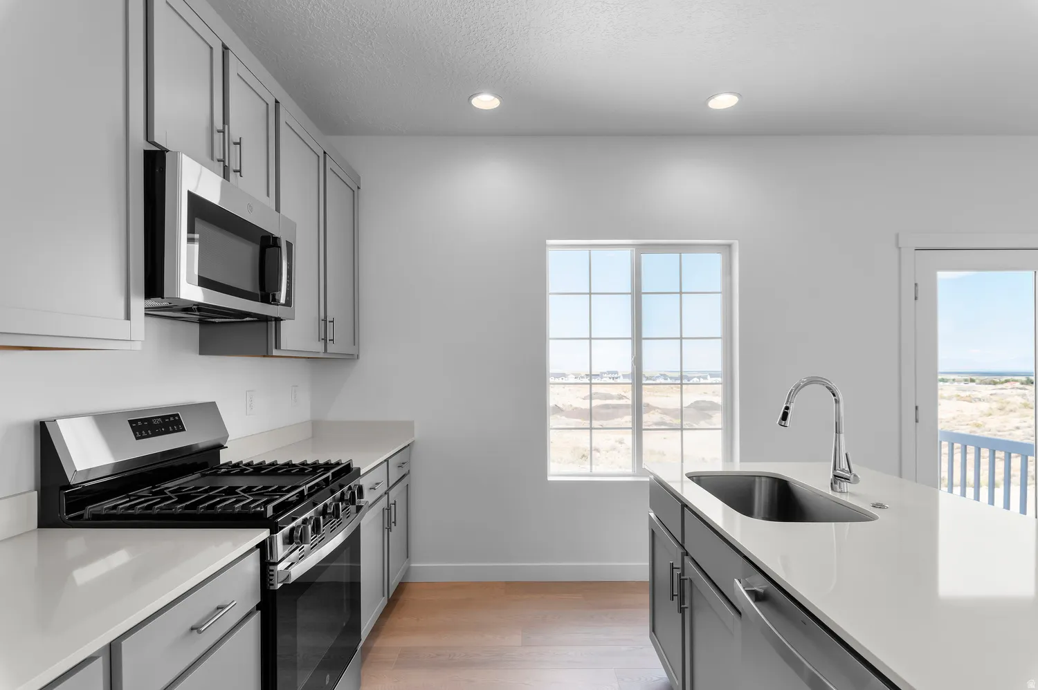 Light gray cabinetry with stainless steel appliances, white countertops, a central island with an undermount sink, and wood-finish flooring