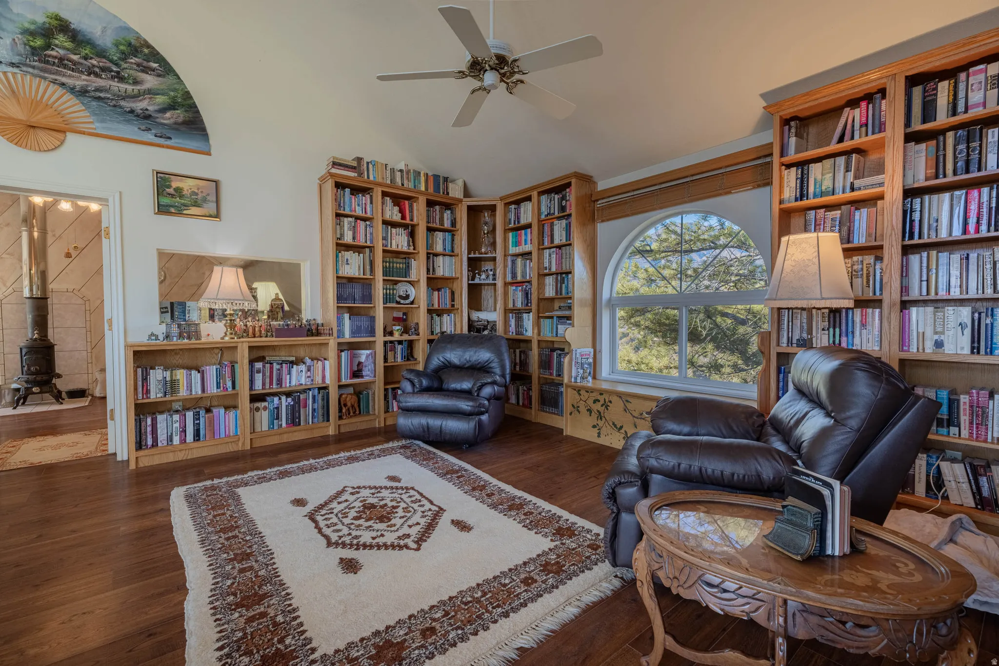 Sophisticated library retreat showcasing extensive built-in wood cabinetry with integrated bookshelves spanning multiple walls, hardwood flooring throughout, arched transom window with natural light, and ceiling fan with brushed-metal finish