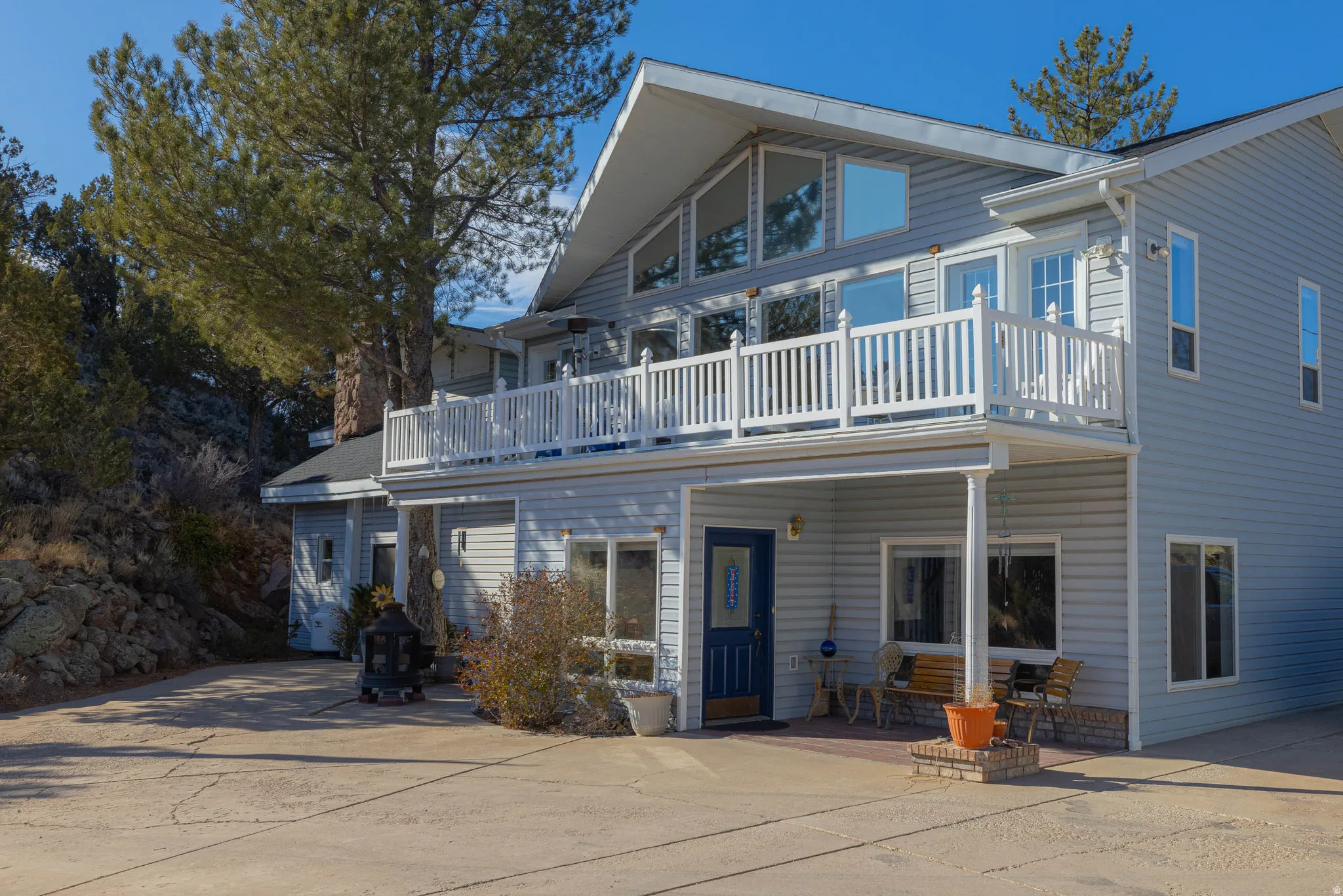 Two-story exterior featuring light blue siding, a prominent second-story balcony with white railing, a ground-level patio, and multiple large windows