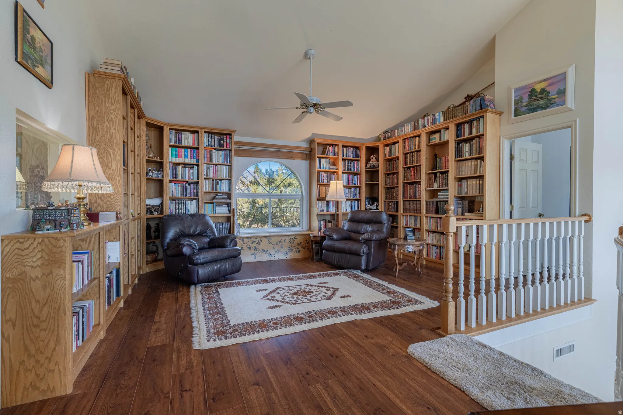 Vaulted ceiling over wood-finish flooring, featuring extensive built-in wooden shelving, an arched window, and a ceiling fan