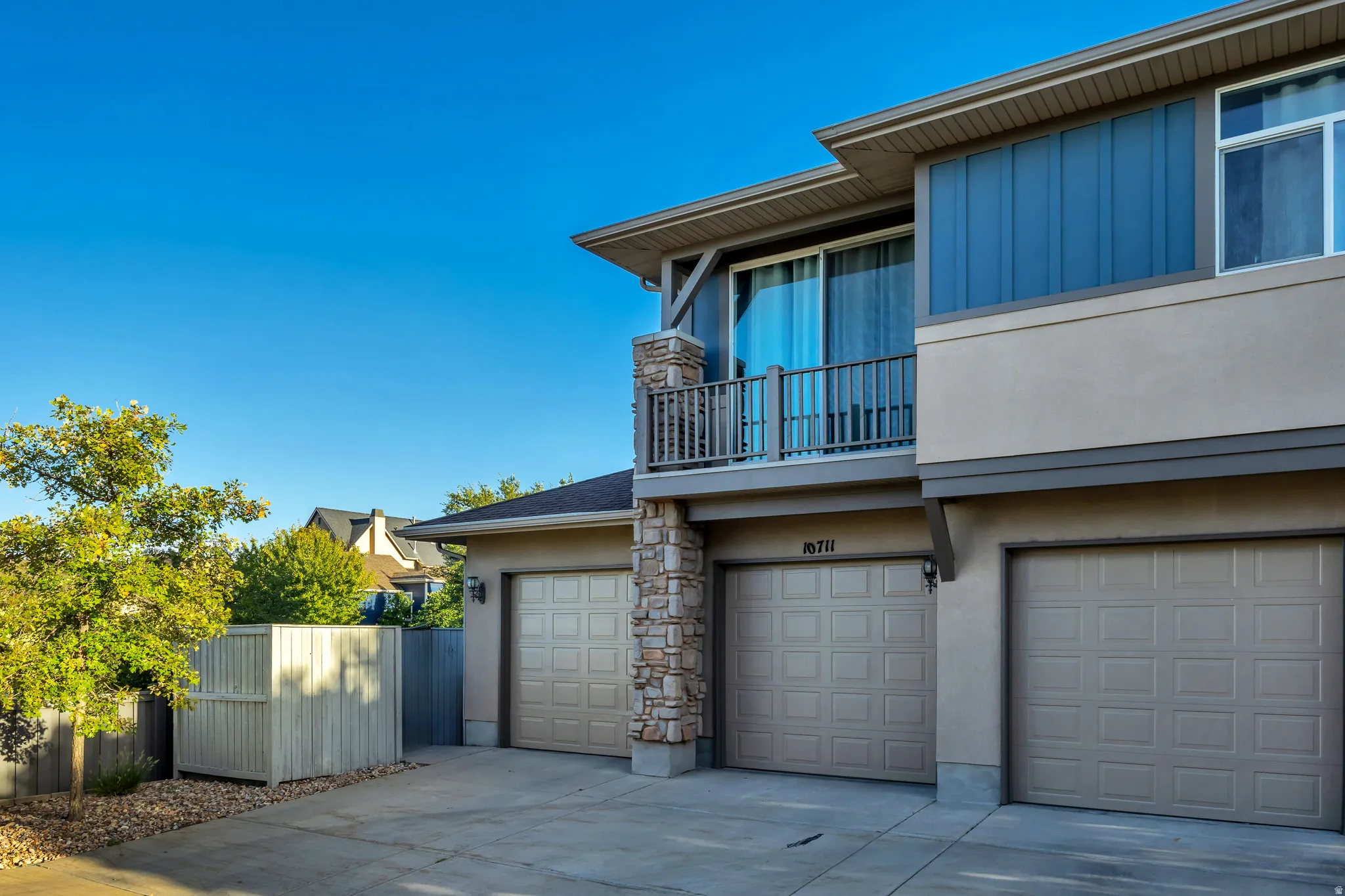 Multi-level exterior with stucco and vertical siding, featuring a private balcony with railing, a stone-clad column, and two garage doors with paneling