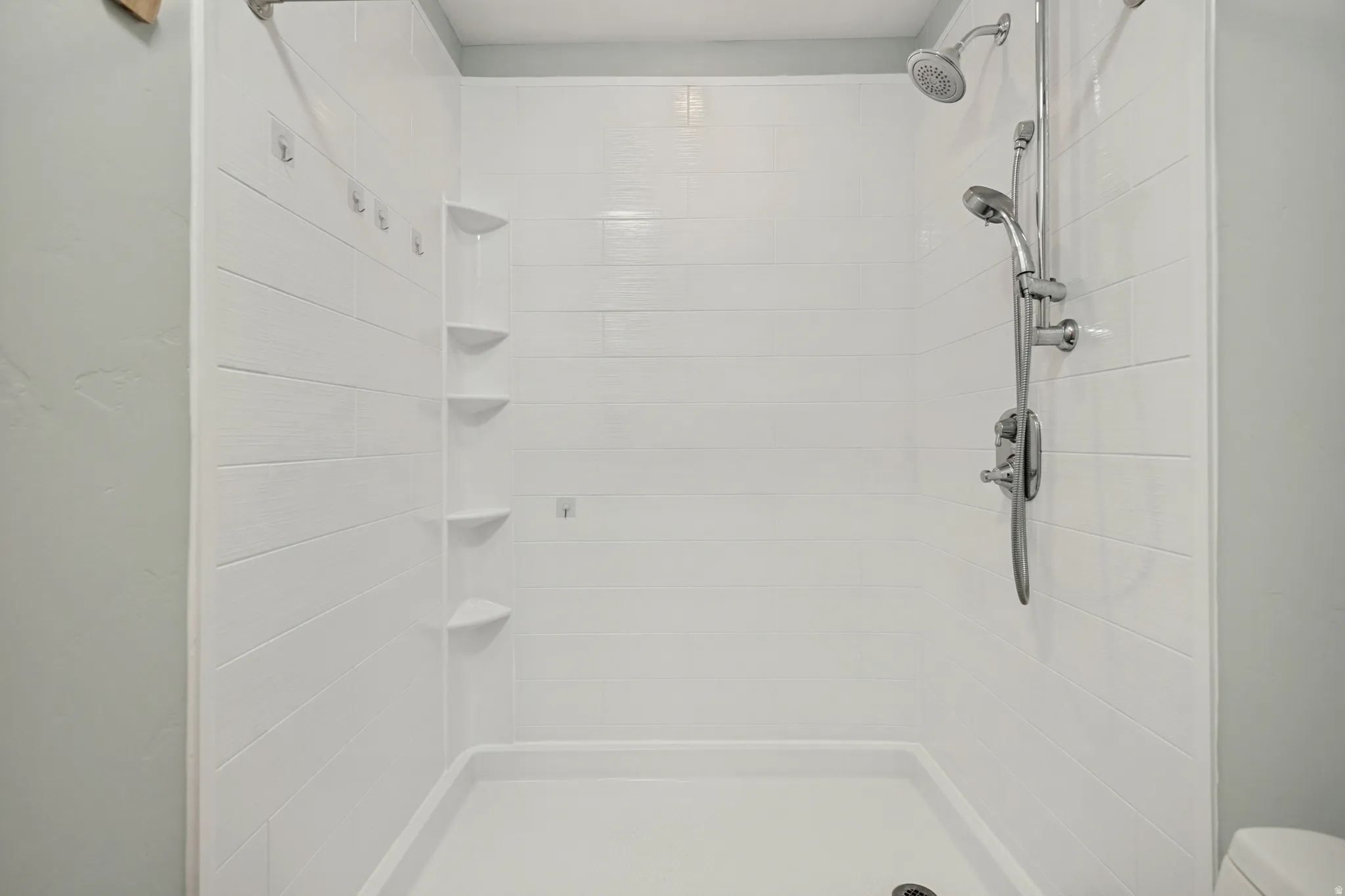 White shower enclosure with a fixed showerhead and a handheld shower attachment, integrated corner shelving, and a textured wall finish