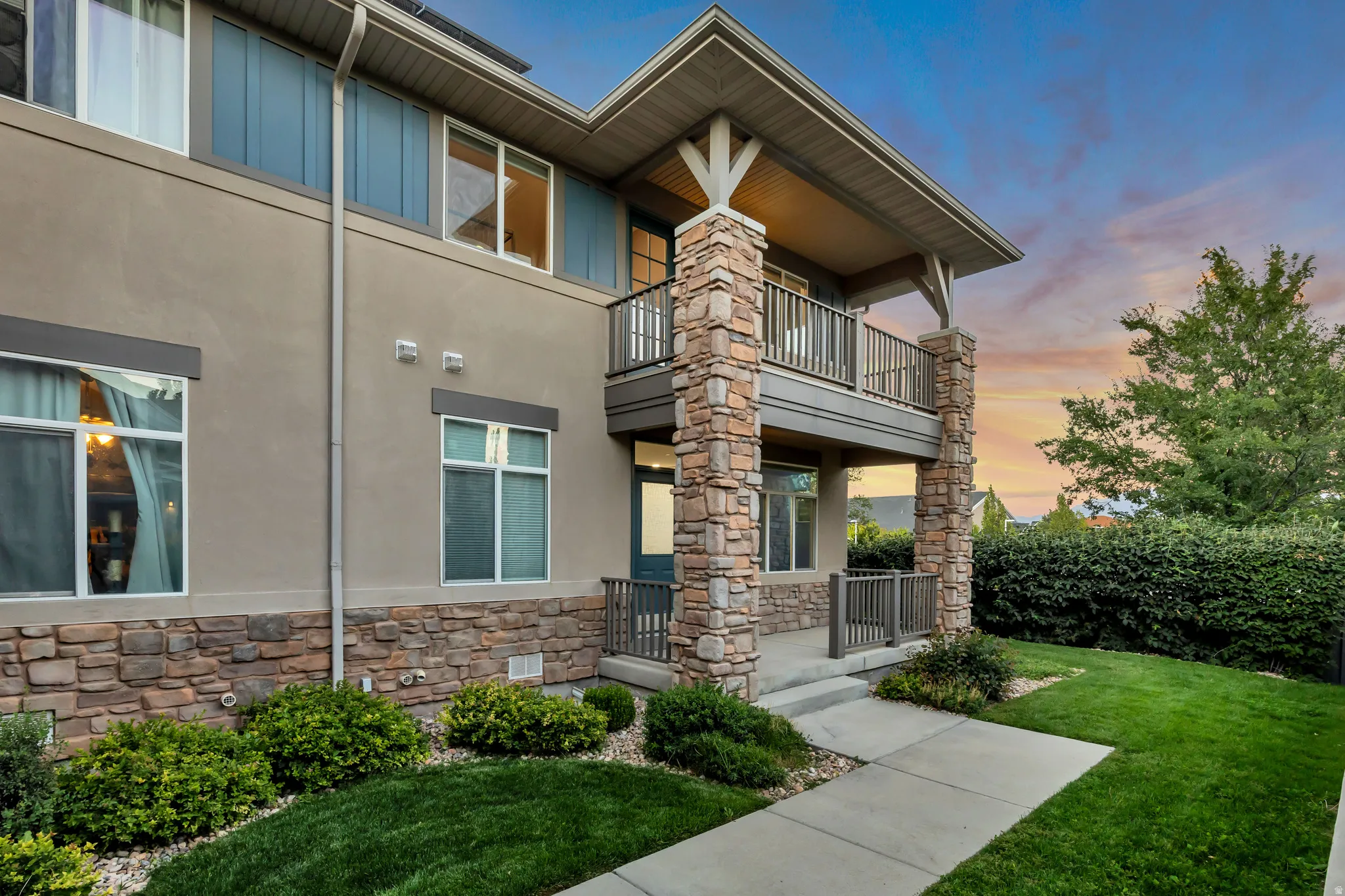 Exterior featuring a stucco facade, stone veneer accents, an upper-level balcony, a ground-level covered entry, and a manicured lawn with shrubbery