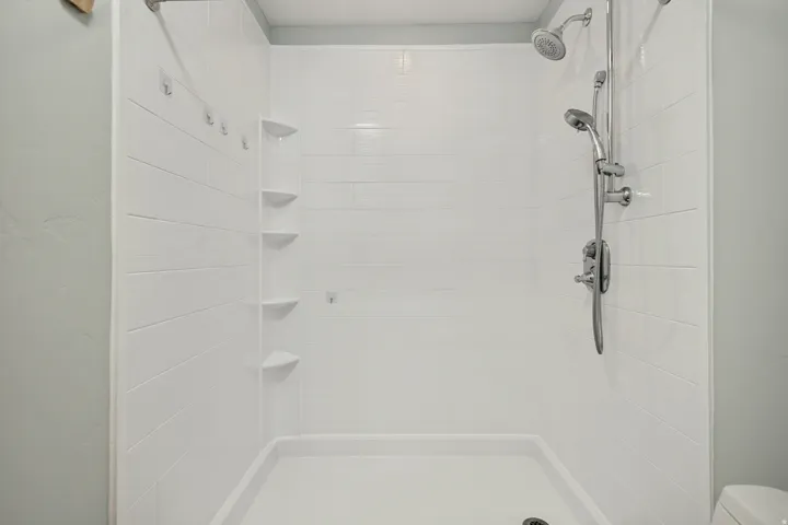 White shower enclosure with a fixed showerhead and a handheld shower attachment, integrated corner shelving, and a textured wall finish