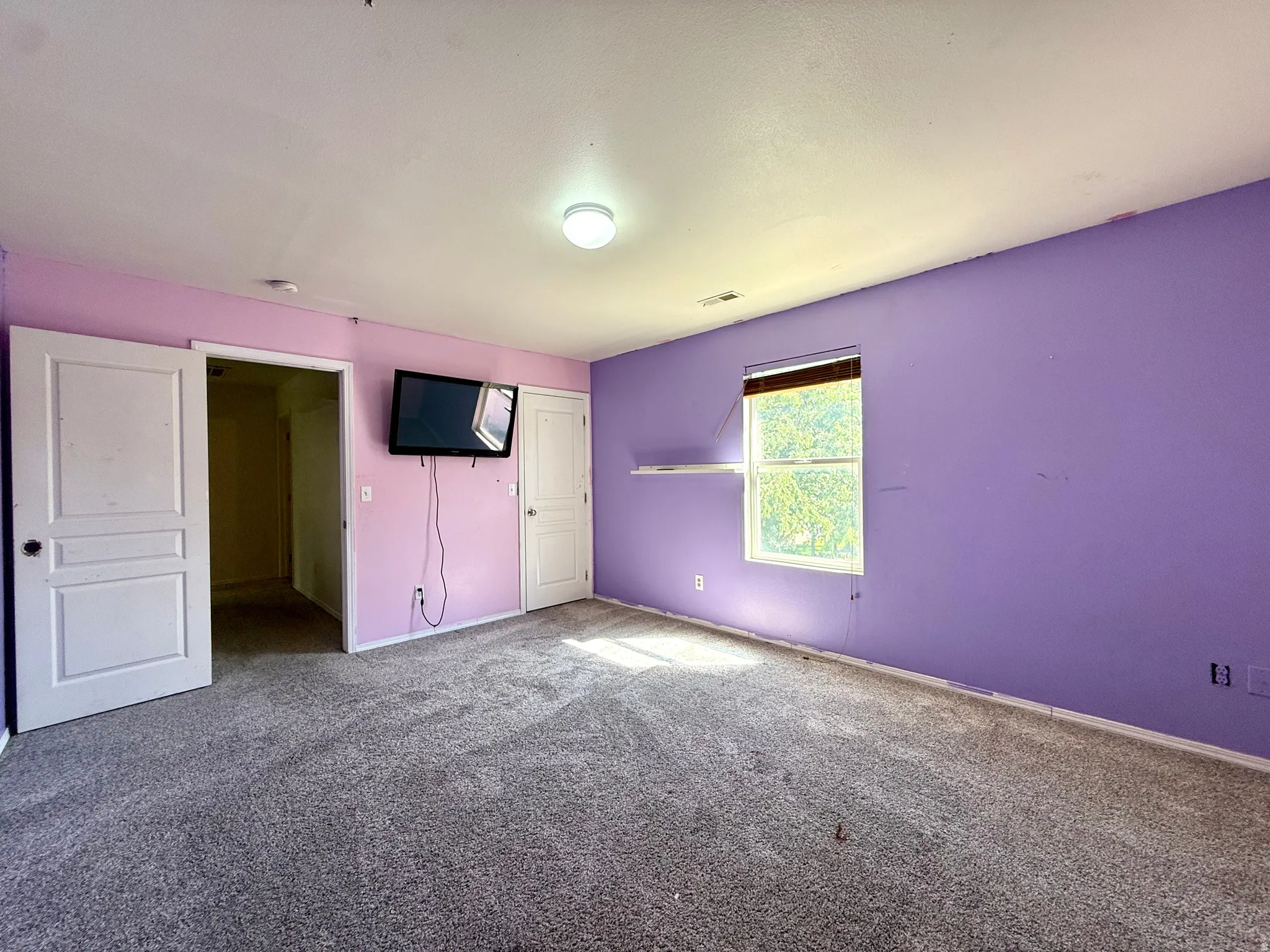 Wall-mounted television bracket, window with blinds, overhead flush-mount light fixture, and textured carpeting