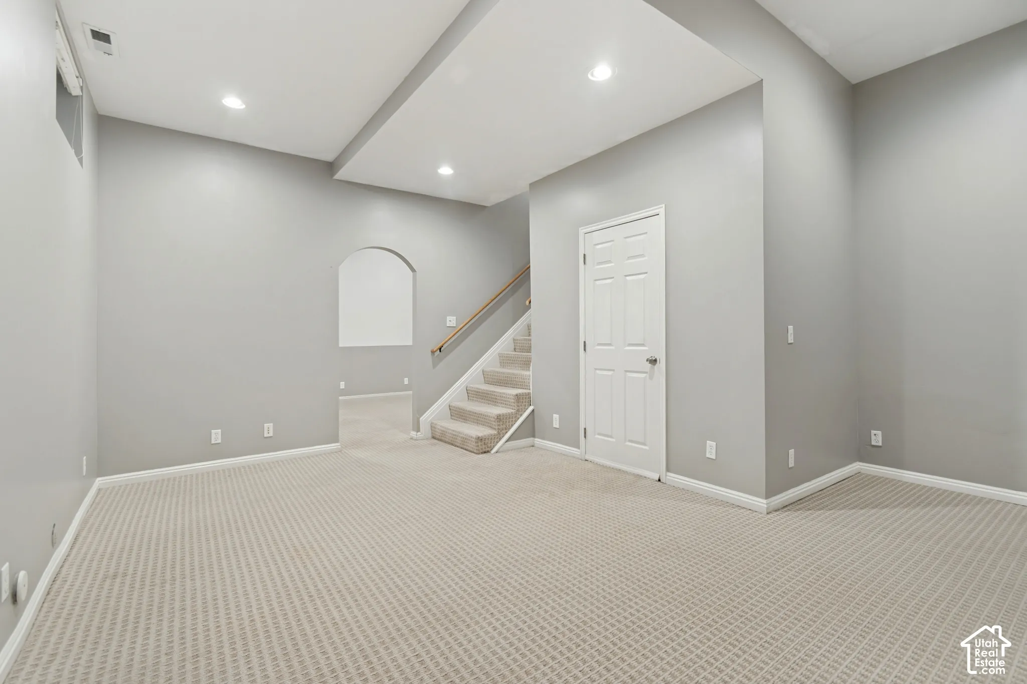 Neutral-toned interior space featuring recessed ceiling lighting, a recessed ceiling tray, wall-to-wall carpeting, an arched entryway, and a staircase with a wooden handrail