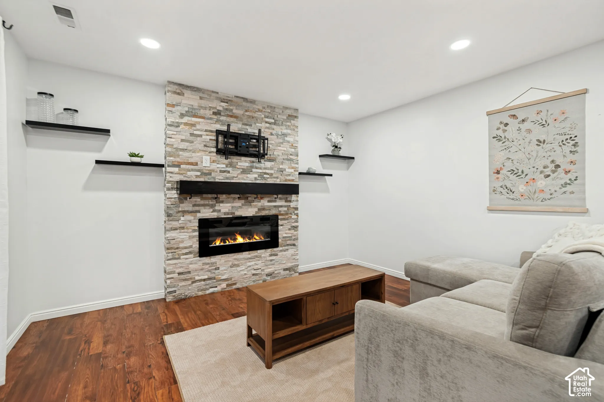 Stacked stone fireplace surround, inset electric fireplace, matte black floating mantel, recessed lighting, and wood-finish flooring