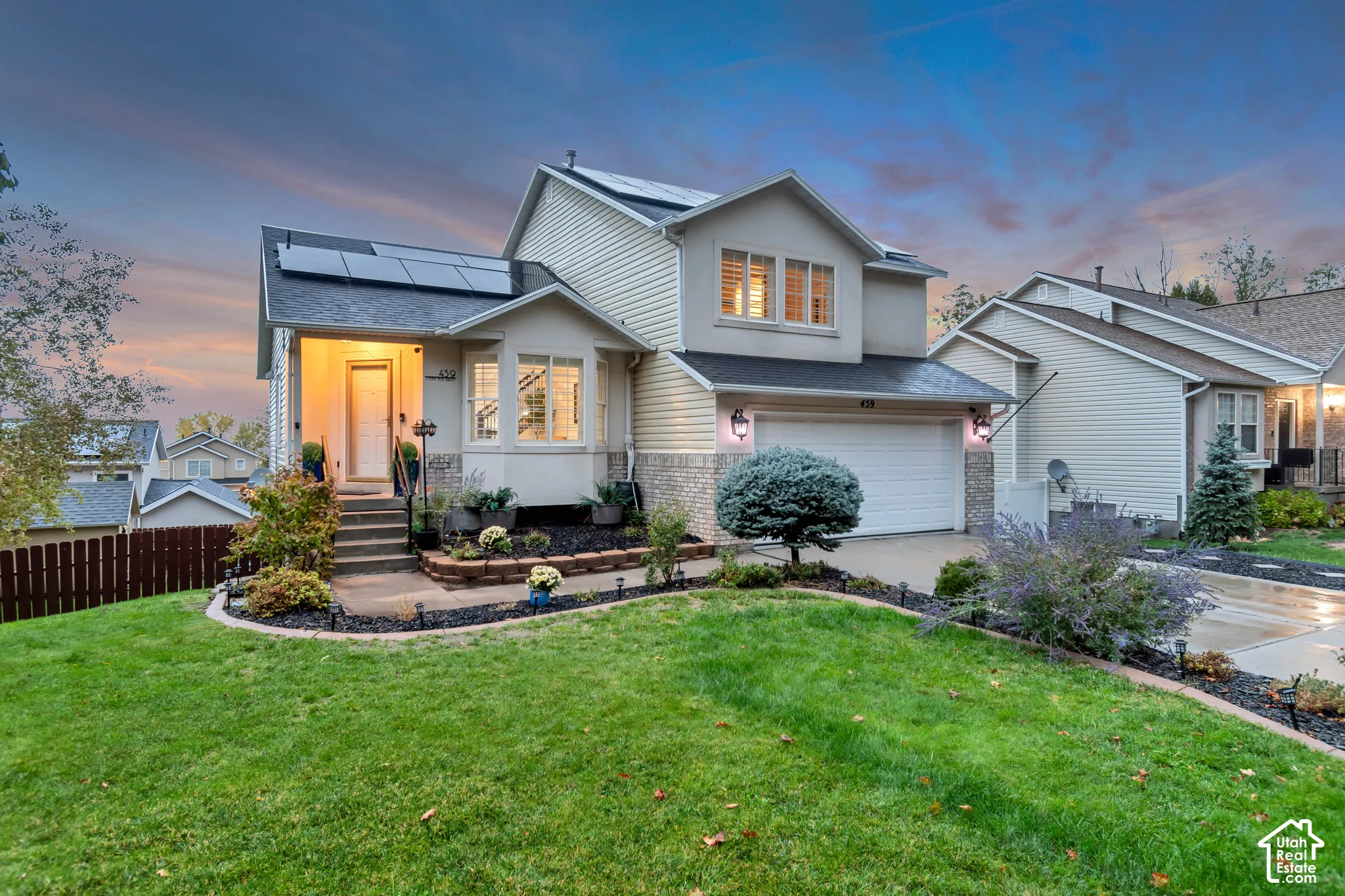 Two-story residence featuring horizontal siding, a two-car garage, roof-mounted solar panels, a covered entryway with stone-veneer accents, and a landscaped front yard with stone-bordered garden beds